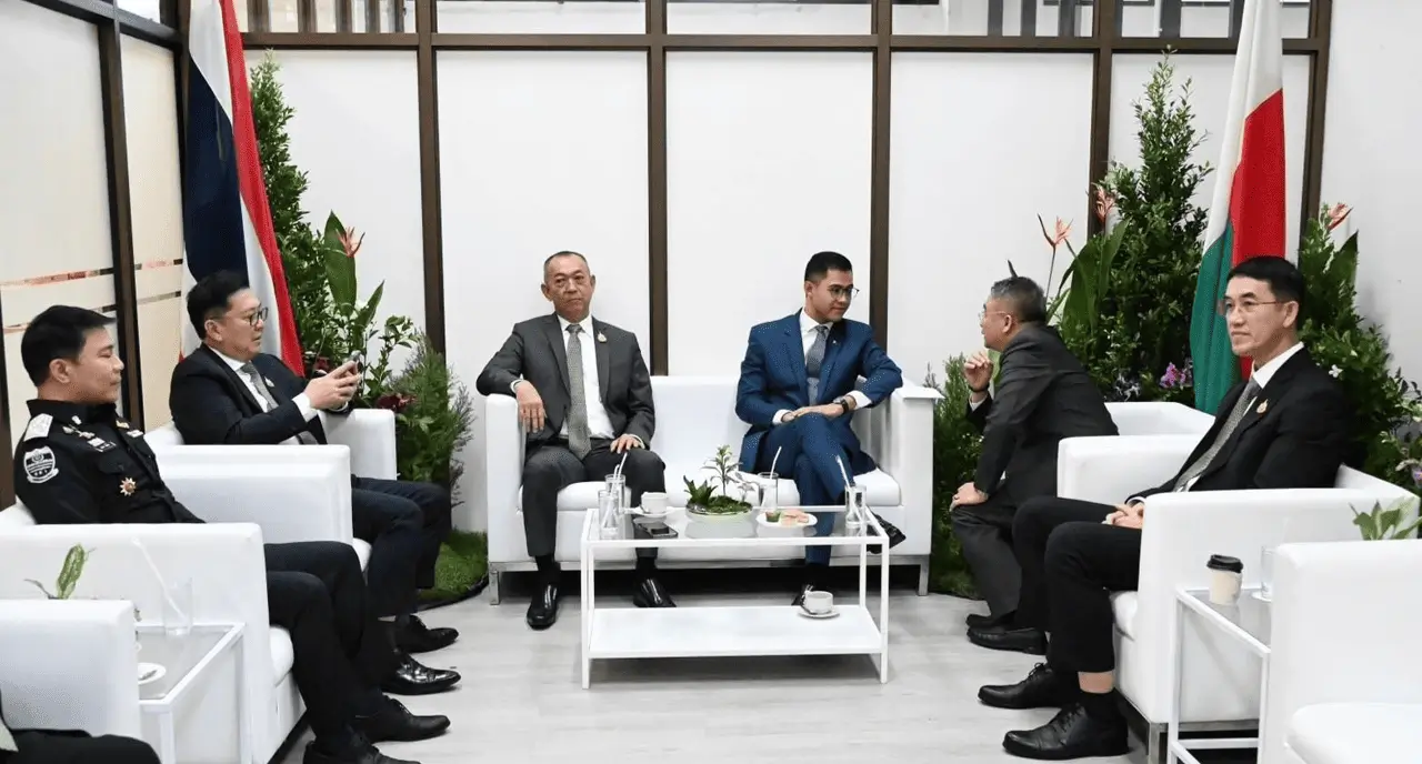 ผู้ชายใส่สูทนั่งคุยกันบนโซฟาสีขาว มีธงชาติประเทศไทย และธงชาติต่างประเทศ