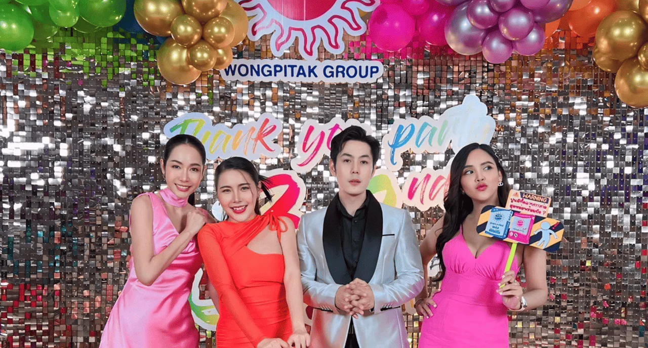 ผู้ชายใส่สูทสีเทายินตรงกลางมีผู้หญิงสามคนใส่ชุดสีชมพูสีแดงและสีชมพูอ่อนกำลังยิ้มให้คนถ่ายภาพที่หน้าแบคดรอปงานเลี้ยง