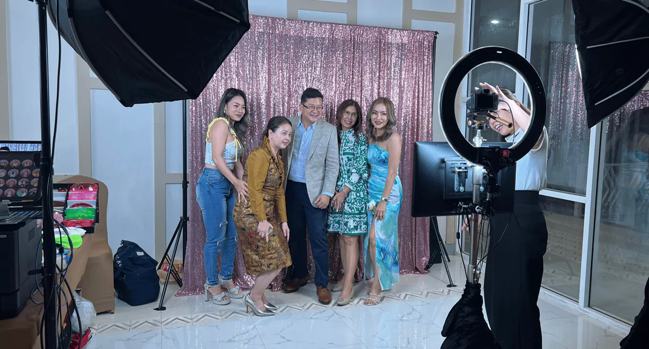 ผู้ชายยืนอยู่ตรงกลางมีผู้หญิงสี่คนกำลังถ่ายรูปภาพร่วมกับผู้ชายด้านหลังเป็นแบคดรอปสีชมพูเลื่อม