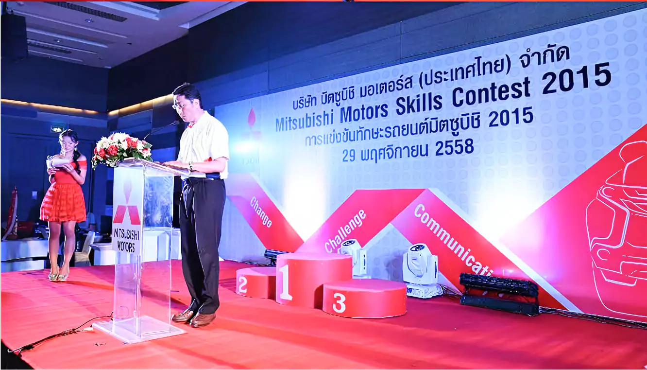 ผู้ชายใส่แว่นยืนอยู่บนเวทีกำลังพูกใส่ไมโครโฟนด้านหน้ามีโพเดียมวางอยู่