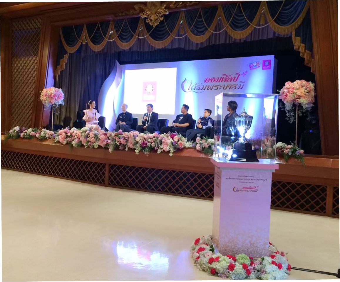 ผู้ชายใส่สูทกำลังถือไมค์นั่งพูดคุยอยู่บนเวที